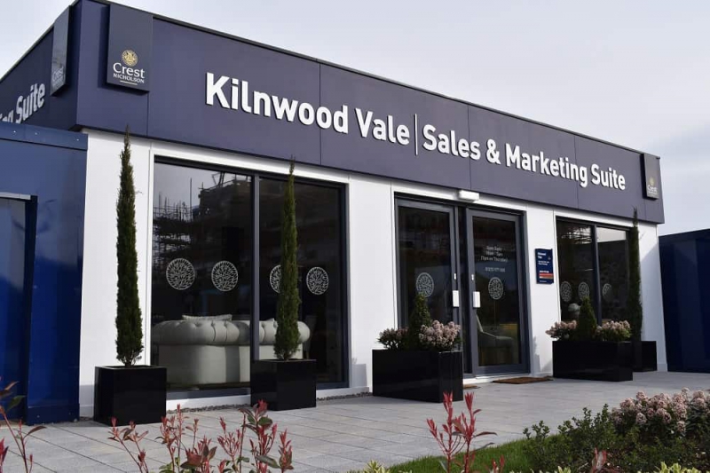 Building's entrance displays text "Kilnwood Vale Sales & Marketing Suite," flanked by two tall potted plants. Inside, white sofas and decorative elements. Exterior includes various plants and tiled flooring.