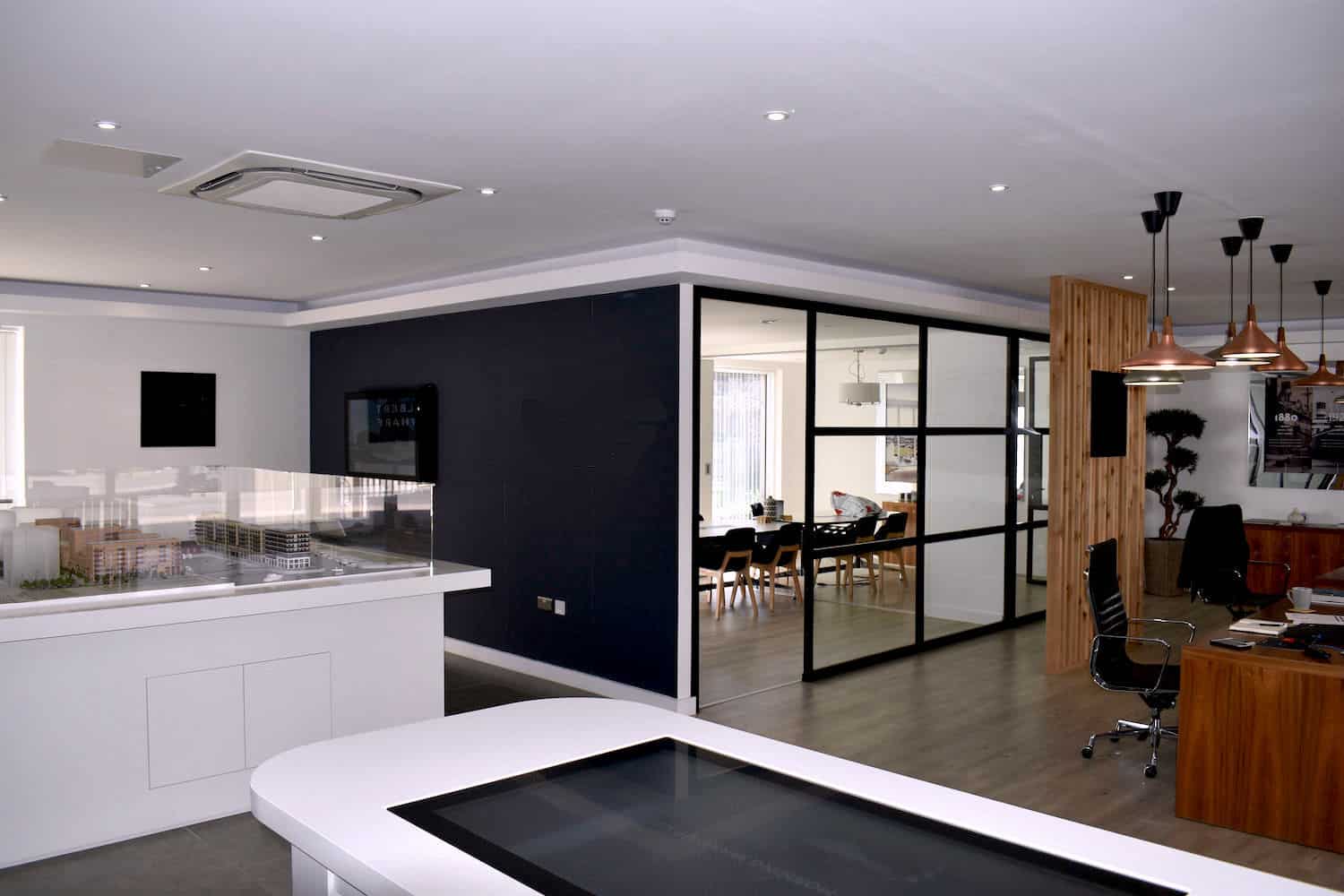 Model of a building sits on a table; conference room with chairs visible through glass wall; modern office setting with wooden accents and pendant lights.