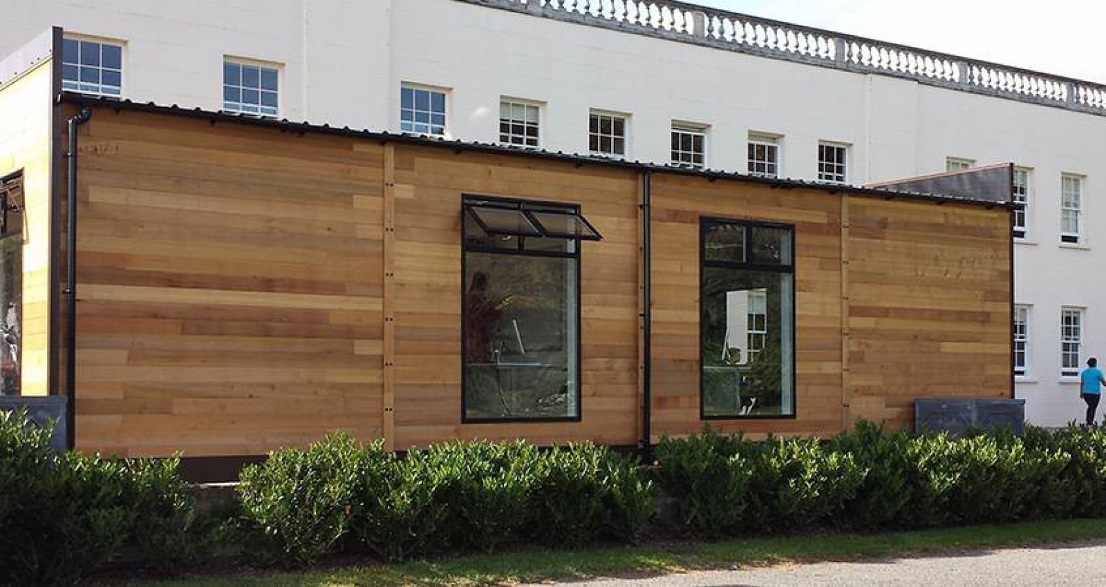 A modern wooden building with large windows sits against an older white structure with several windows, surrounded by greenery on a clear, sunny day.