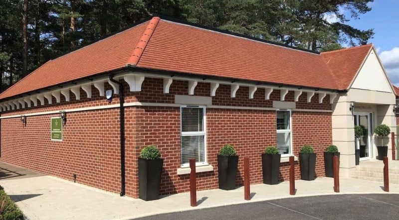 A small brick building with a red-tiled roof, windows lined with tall planters holding bushes, situated in a wooded area. Sign text: "Welcome to Wellington Cemetery."