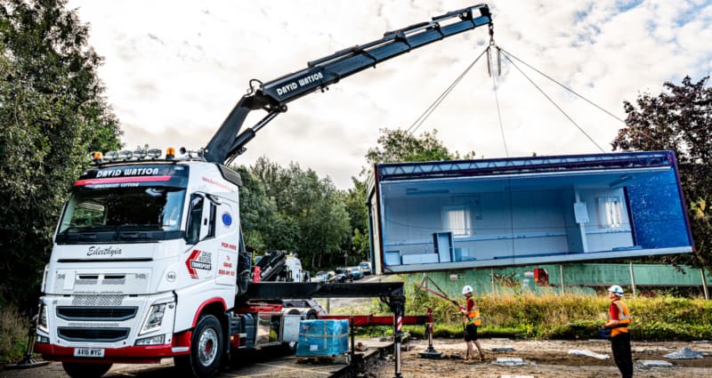 A truck with "David Watson" branding uses a crane to lift a large container in a grassy, industrial area, while two construction workers in safety gear observe.