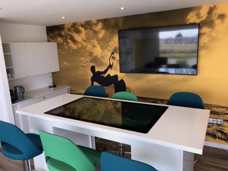 A white table surrounded by colorful chairs has a large monitor on the wall with a silhouette of a kiteboarder and a dramatic sunset scene in the background.