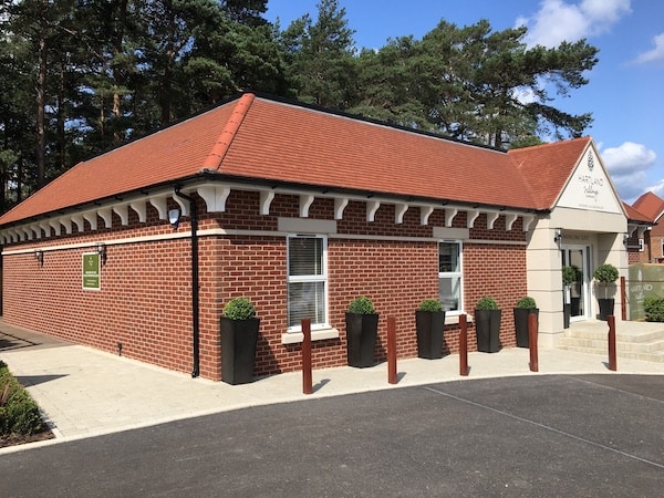 A single-story brick building with a red-tiled roof stands surrounded by a paved sidewalk and tall, black planters with greenery. Pine trees are visible in the background.