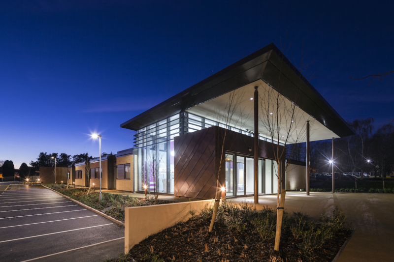 A modern building with large glass windows, illuminated at night, stands in a landscaped area next to a nearly empty parking lot, surrounded by trees and streetlights.