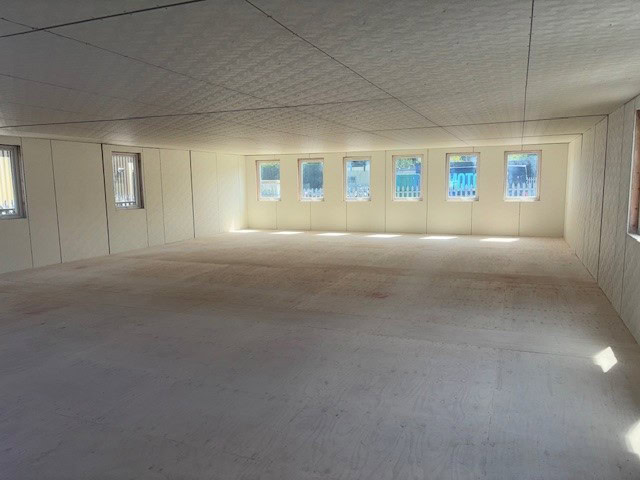 An empty, spacious, white-walled room with a wooden floor and several windows letting in natural light; outside scenery is partially visible through the windows.