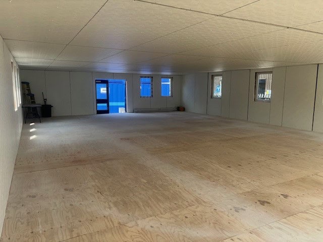 A spacious, unfinished room with a plywood floor and several small windows along the walls; one side features a blue door and scattered objects like tables and bins.