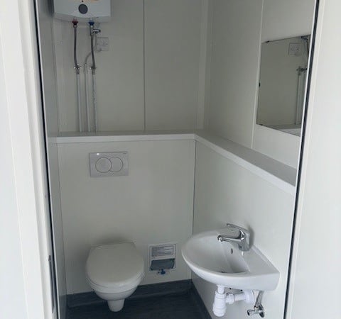 A small, compact bathroom features a wall-mounted toilet, a sink with a faucet and drainpipe, a mirror above the sink, and a water heater with exposed pipes on the wall.