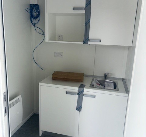 A small kitchenette with white cabinets, a sink, and blue cables on the left wall; the room appears newly installed with protective tapes on the cabinets.