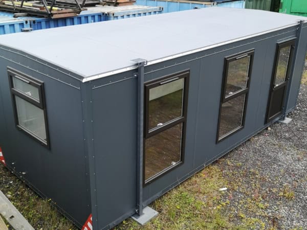A dark grey rectangular portable building stands on a gravel and grassy area. It has multiple rectangular windows, a door, and containers stacked nearby.
