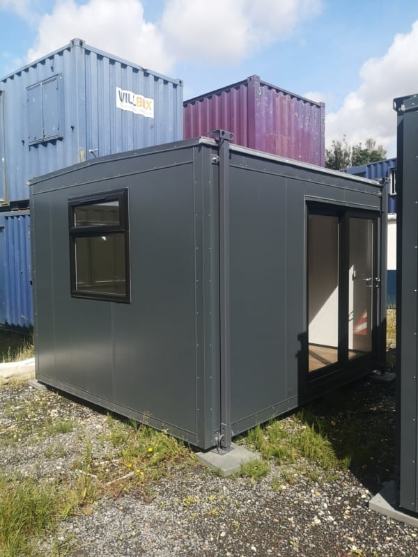 A gray metal storage container with a window and sliding glass door stands on uneven ground with weeds, surrounded by stacked blue and purple shipping containers. "VILIBIX" is text on a blue container.