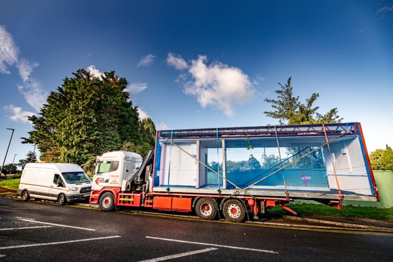 A red and white truck transports a large glass structure on its flatbed trailer, parked on a suburban street next to a white van with trees in the background.